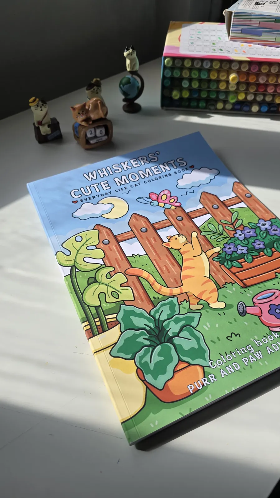 The coloring book on a sunny desk next to a box of markers and small cat figurines